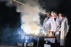 Two people in white lab coats stand intently at a table covered with various laboratory equipment and chemicals. Thick clouds of smoke rise from the center of the table, illuminating the dark background.