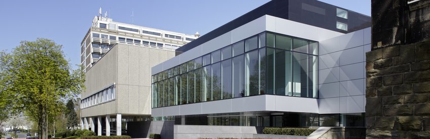 Administration Modern building with glass façade, adjacent to a beige concrete structure, surrounded by trees.