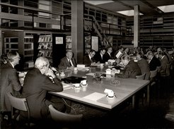 Eine Gruppe von Männern sitzt um einen großen Tisch in einem Büro, mit zahlreichen Bücherregalen und Dokumenten auf dem Tisch. Tassen und Aschenbecher sind ebenfalls sichtbar.
